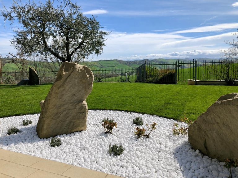 Modern landscape garden with large boulders in white gravel, green lawn, oak tree, and countryside views behind black fence