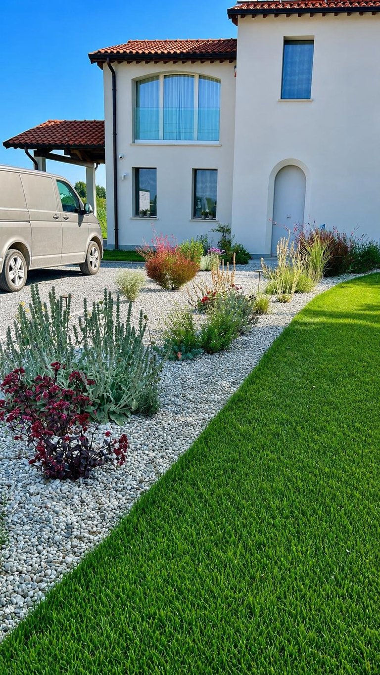 Modern Mediterranean-style house with white stucco exterior, red tile roof, green lawn, and decorative gravel landscaping with ornamental plants and a parked van.
